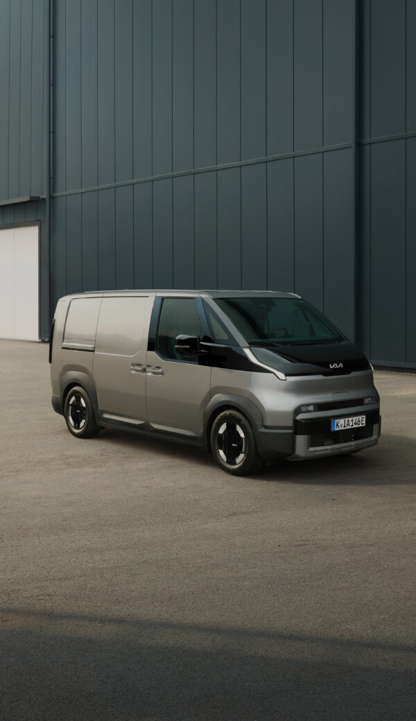 Heckansicht des Kia PV5 Cargo, angeschlossen an eine Ladestation, mit einem Mann, der daneben an einem Laptop arbeitet.